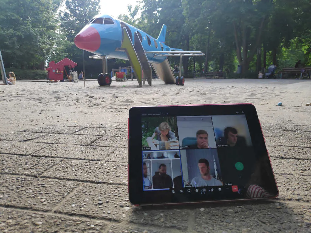 Im Vordergrund Tablet mit Meeting Video der Generalversammlung, im Hintergrund Spielplatz mit Kletterrakete